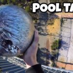 Person grasps a bowling ball from a high balcony, looking down on a courtyard with a small pool table and the words 'POOL TABLE' marked by an arrow.