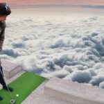 Man in a casual sweatshirt and dark pants stands on a small green practice mat on a rooftop, preparing a golf swing with clouds below at sunset.